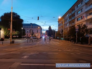 Zdjęcie kierujących ruchem dwójki policjantów wrocławskiej drogówki na jednej z wrocławskich ulic. W tle policyjne radiowozy z włączonymi sygnałami świetlnymi.