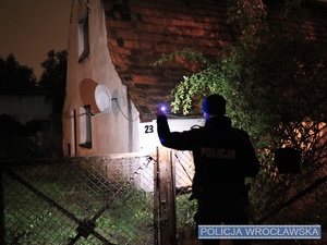 Zdjęcie stojącego przy ogrodzeniu opuszczonego budynku umundurowanego policjanta oświetlającego latarką na tern posesji