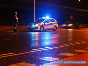 Kierujący nocą ruchem na jednej z wrocławskich ulic umundurowany policjant ruchu drogowego stojący przy oznakowanym policyjnym radiowozie.