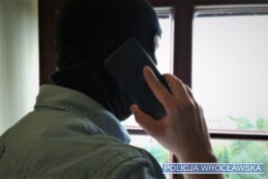 Mężczyzna w kominiarce trzyma w dłoni telefon, stoi na tle okna