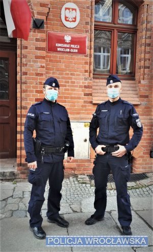 Zdjęcie przedstawia dwóch policjantów w granatowych mundurach oraz maseczkach na twarzy, którzy stoją przed ceglanym budynkiem. Na budynku umieszczona jest czerwona tablica z białym napisem Komisariat Policji Wrocław-Ołbin