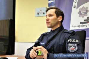 Policjant w granatowym mundurze, prowadzący spotkanie online, siedzący przy biurku na tle logo Komendy Miejskiej Policji we Wrocławiu