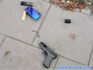 Przedmiot wyglądający jak pistolet, telefon komórkowy oraz zapalniczka leżące na chodniku