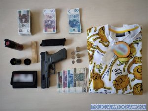 Od lewej portfel, gotówka, pojemnik z gazem, przedmiot przypominający broń, monety oraz biała bluzka w żółte wzory