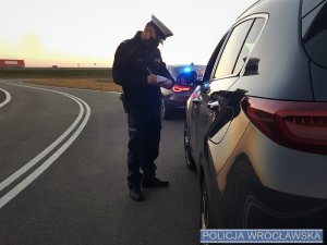 Umundurowany policjant w białej czapce stojący obok czarnego auta