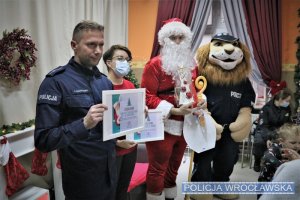 Dzieci wręczają podziękowania. Od lewej: policjant, przedstawicielka centrum handlowego, Święty Mikołaj oraz Komisarz Lew