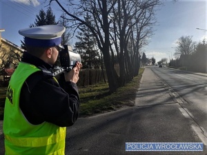 Zdjęcie poglądowe umundurowanego policjanta w trakcie wykonywania pomiaru prędkości przy pomocy ręcznego miernika prędkości
