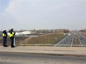 Umundurowany funkcjonariusz w trakcie wykonywania pomiaru odstępów między pojazdami ręcznym miernikiem stojący na wiadukcie nad drogą wielojezdniową