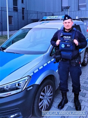 Umundurowany policjant stojący obok oznakowanego radiowozu na tle budynku