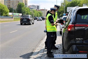 Dwoje umundurowanych funkcjonariuszy w trakcie kontroli kierującego pojazdem stojący na drodze przy aucie osobowym