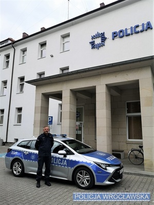 Umundurowany policjant stojący przy oznakowanym radiowozie na tle budynku komisariatu