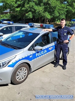 Umundurowany policjant stojący przy oznakowanym radiowozie na placu