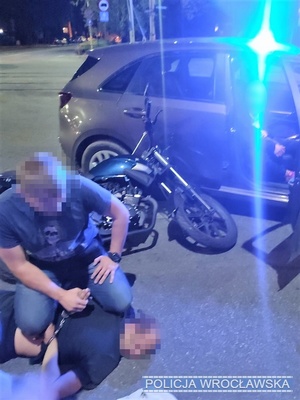 Na zdjęciu widać zatrzymanego przez nieumundurowanych policjantów mężczyznę a w tle motocykl i nieoznakowany radiowóz z włączonymi sygnałami świetlnymi