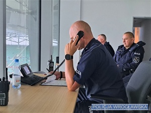 Siedzący przy biurku, w pomieszczeniu, umundurowany policjant odbierający telefon oraz siedzący za nim dwaj umundurowani policjanci