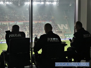 Siedzący przy biurku umundurowani policjanci na tle widoku na boisko piłkarskie i trybuny