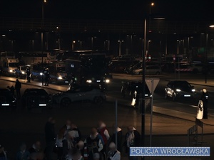 Grupa osób stojących na parkingu na tle oznakowanych radiowozów