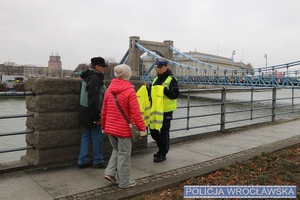 Zdjęcie poglądowe - umundurowana policjantka Wydziału Ruchu Drogowego rozdająca na skwerze przy Moście Grunwaldzkim kamizelki odblaskowe dwóm osobom.