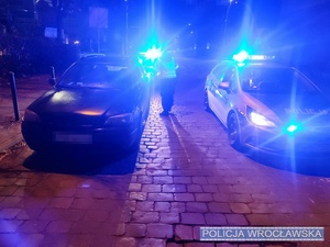 Zdjęcie stojącego nocą osobowego auta a obok oznakowanego radiowozu Policji z włączonymi światłami uprzywilejowania na jednej z wrocławskich ulic