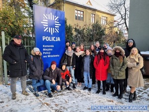 Grupa studentów wraz z wykładowca w tle baner reklamowy Komendy Miejskiej Policji we Wrocławiu