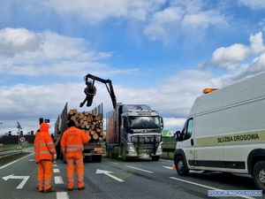 Zdjęcie pojazdu ciężarowego stojącego na jednej z podwrocławskich dróg z rozsypanym ładunkiem drewna leżącym na jezdni i pojazdem ponownie ładującym go na pojazd oraz służbą drogową zabezpieczająca miejsce zdarzenia