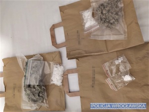 Leżące na stole torby papierowe oraz ponumerowane torby foliowe zawierające różnego rodzaju substancje, w tym susz roślinny, proszki, kryształki i tabletki