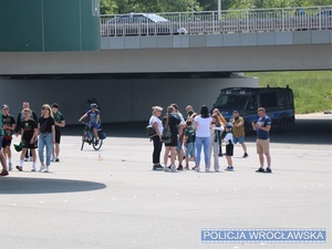 Idący placem obok stadionu kibice oraz stojący w tle oznakowany radiowóz Policji