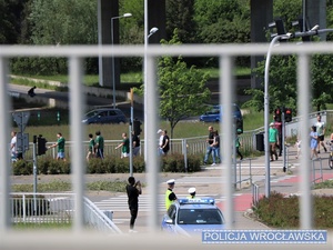 Idący ulicą obok stadionu kibice oraz stojący w tle oznakowany radiowóz Policji i dwaj policjanci Wydziału Ruchu Drogowego