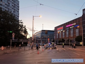 Stojący na jednej z wrocławskich ulic oznakowany radiowóz policji z włączonymi sygnałami świetlnymi pojazdu uprzywilejowanego oraz umundurowany funkcjonariusz kierujący ruchem