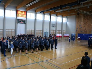 Policjanci stojący na sali w mundurach galowych podczas uroczystości z okazji Święta Policji w tle zaproszeni goście oraz prowadzący uroczyści i lektor