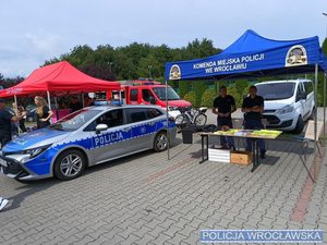 Policyjny radiowóz w tle policjanci w mundurze na stoisku prewencyjnym oraz zainteresowani przechodnie