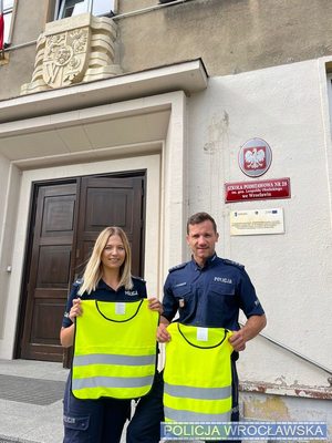 policjanci w mundurach przed szkołą podstawową trzymają w rękach odblaskowe kamiezelki