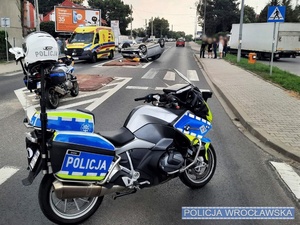 Zdjęcie stojącego na drodze oznakowanego policyjnego motocykla z widocznym w tle pojazdem, który brał udział w zdarzeniu drogowym leżącym na dachu