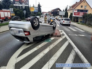 Zdjęcie stojących na drodze oznakowanych policyjnych motocykli z widocznym na pierwszym tle pojazdem, który brał udział w zdarzeniu drogowym leżący na dachu