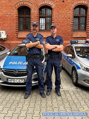 Dwaj umundurowani policjanci stojący na tle zaparkowanych radiowozów i ceglastego budynku