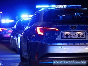 Zdjęcie stojących nocą, na jednej z wrocławskich ulic, jeden z drugim trzech oznakowanych radiowozów Policji z włączonymi sygnałami świetlnymi pojazdu uprzywilejowanego