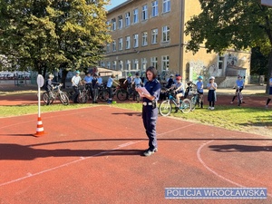 Stojąca na płycie boiska szkolnego jednej z wrocławskich szkół umundurowana policjantka wraz z uczniami szkoły przygotowanymi do egzaminu na kartę rowerową.