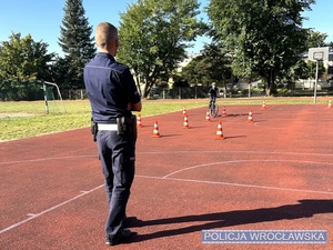 Stojący na płycie boiska szkolnego jednej z wrocławskich szkół umundurowany policjant wraz z uczniami szkoły przygotowanymi do egzaminu na kartę rowerową.