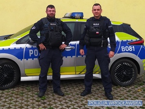 Dwaj umundurowani policjanci stojący na tle oznakowanego radiowozu Policji.