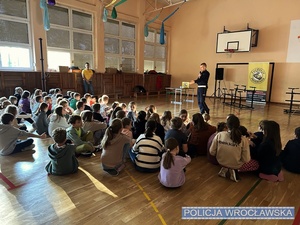 Zdjęcie, na którym widać prowadzącego, na sali gimnastycznej, spotkanie z dziećmi funkcjonariusza Policji dotyczące bezpieczeństwa w ruchu drogowym.