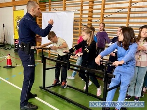 Stojący na sali gimnastycznej uczniowie szkoły podstawowej oraz stojący przed nimi umundurowany policjant prowadzący zajęcia z bezpieczeństwa w ruchu drogowych, m.in. symulatorze jazdy na rowerze.