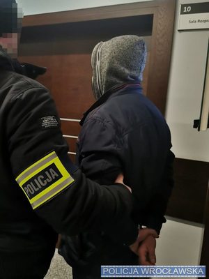 policjant w ubraniu cywilnym obok zatrzymany mężczyzna z rękami w kajdnkach z tyłu stoją przy sali sądowej