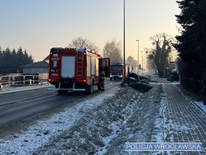 Zdjęcie z miejsca zdarzenia drogowego na drodze w jednej z podwrocławskich miejscowości gdzie pojazd osobowy marki Peugeot uderzył przydrożną latarnie, a w tle widać funkcjonariuszy straży pożarnej oraz ich pojazdy.