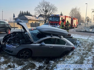 Zdjęcie z miejsca zdarzenia drogowego na drodze w jednej z podwrocławskich miejscowości gdzie pojazd osobowy marki Peugeot uderzył przydrożną latarnie, a w tle widać funkcjonariuszy straży pożarnej oraz ich pojazdy.