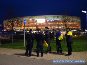 Umundurowani policjanci prewencji stojący na chodniku przed stadionem i rozmawiający z kibicami udającymi się na mecz piłkarski.