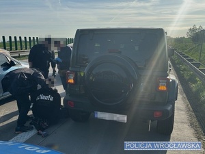 Stojący na autostradzie samochód marki Jeep oraz stojący przy nim funkcjonariusze policji, w tym mundurowi z ruchu drogowego oraz siedzący na jezdni zatrzymany mężczyzna.