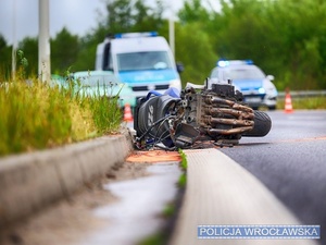 Leżący na jezdni motocykl biorący udział w wypadku na tle oznakowanych radiowozów Policji.