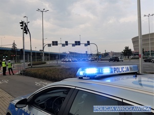 Zdjęcie umundurowanych funkcjonariuszy ruchu drogowego stojących przy oznakowanym przejściu dla pieszych obok oznakowanego radiowozu w trakcie wykonywania czynności związanych z nadzorowaniem ruchu drogowego.