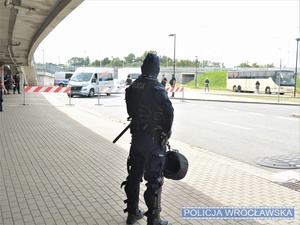 Zdjęcie umundurowanego funkcjonariusza Oddziałów Prewencji Policji w trakcie wykonywania czynności związanych z nadzorowaniem przejścia kibiców przy stadionie.