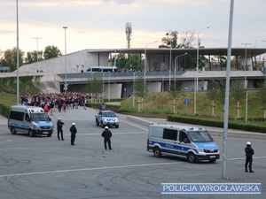 Wchodzący na stadion kibice w asyście umundurowanych policjantów Oddziałów Prewencji Policji oraz stojących obok oznakowanych radiowozów.