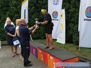 Komendant Miejski Policji we Wrocławiu mł. insp. Rafał Siczek wręczający puchar stojącemu na podium jednej z uczestniczek czwartej edycji Mistrzostw Senioralnych o Puchar Komendanta Miejskiego Policji.